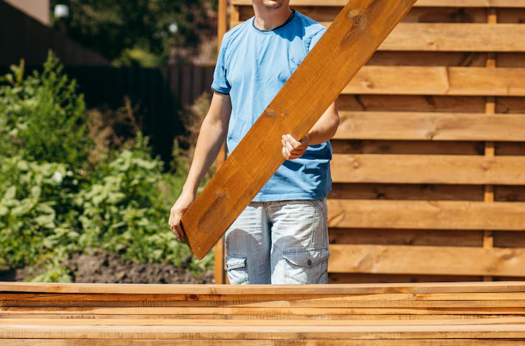 Man holding wooden plank for construction in garden, standing next to wooden fence wearing blue t-shirt and shorts